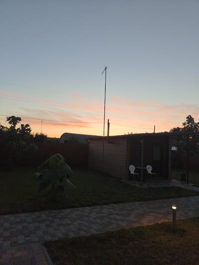 This media file depicts a serene outdoor scene at dusk in Shkurupiivka, Ukraine. The sky is a gradient of soft blues and pinks, with wispy clouds illuminated by the setting sun. A tall antenna stands prominently against the twilight sky.
In the foreground, a paved pathway made of interlocking gray bricks leads the eye towards a wooden structure. This building appears to be a shed or a small outbuilding, with a covered porch area. Two white chairs are placed on the porch, suggesting a place for relaxation. Adjacent to the pathway, a small outdoor lamp is lit, casting a warm glow on the surrounding grass.
The scene is framed by lush greenery. To the left, a large leafy plant stands out against the darker background. Trees and bushes create a natural border, with a large, arched corrugated metal structure visible in the distance behind the wooden building. The overall atmosphere is peaceful and tranquil, indicative of the end of a day. The time of day is clearly sunset, and the weather appears clear. No visible text is present in the image.