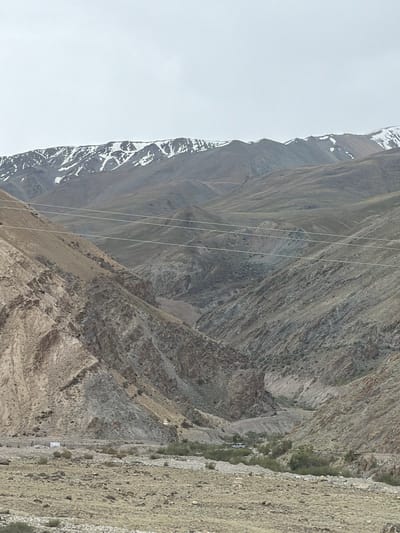 The image shows a rugged mountain landscape with snow-capped peaks in the distance. The mountains are steep and rocky, with some vegetation in the valleys. There are power lines running across the scene. In the foreground, there is a dry, rocky riverbed with sparse vegetation. A small, white structure and a vehicle are visible in the distance. The sky is overcast. This scene is consistent with the high-altitude environment of Stongde, India.