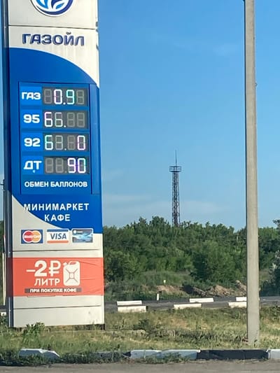 This image captures a roadside view with a gas station price sign as the primary focus. The sign displays prices for different types of fuel: "ГАЗ" (Gas) at 80.98, "95" (likely 95 octane gasoline) at 68.88, "92" (likely 92 octane gasoline) at 68.00, and "ДТ" (Diesel Fuel) at 88.90. Above the fuel prices, it indicates "ОБМЕН БАЛЛОНОВ" (Bottle Exchange) and also advertises a "МИНИМАРКЕТ" (Minimarket) and "КАФЕ" (Cafe). Payment options like Visa and Mastercard are also visible. A special offer is advertised: "-2 РУБЛЯ" (2 Rubles) for a "ЛИТР" (Liter) with the condition "ПРИ ПОКУПКЕ КОФЕ" (When buying coffee). The gas station appears to be branded as "ГАЗОЙЛ".
In the background, a transmission tower or similar structure is visible against a clear blue sky, suggesting it's daytime with fair weather. Green trees and vegetation line the right side of the image, indicating a semi-rural or suburban setting, possibly along a highway. A concrete barrier or curb is also visible. The image seems to be taken from a moving vehicle, as evidenced by the slight blur and the angle of the shot. The overall scene is typical of a gas station encountered during travel.