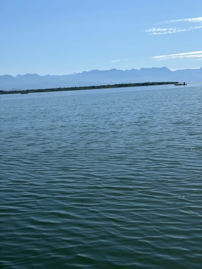 The image captures a broad expanse of water in the city of Braćeni, Montenegro, under a clear, light blue sky that contains minimal wispy white clouds on the right side. A distant horizontal mountain range, rendered in muted bluish-grey tones, occupies the background, partially obscured by atmospheric haze. Immediately below the mountains, a long, dark green landmass, appearing as an island or shoreline, stretches across the frame. The foreground and mid-ground consist of dark blue-green water, characterized by numerous small, parallel ripples and waves reflecting sunlight as scattered glints, particularly pronounced on the right. On the water in the mid-distance, two slender, dark-colored boats are visible. The boat on the far right features an outboard motor and a discernible dark figure, positioned as if operating the vessel and moving towards the right. A second, similar boat is situated further to the left, closer to the center of the frame, near the distant landmass. Several indistinct small dark specks are scattered on the water's surface. The scene is illuminated by bright daylight.
