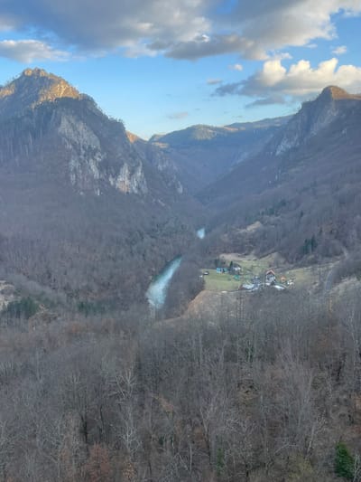 The image presents an expansive aerial view of the Tara River Canyon near Đurđevića Tara, Montenegro. The scene is dominated by rugged, steep mountainsides flanking a deep valley. These slopes are extensively covered in dense, bare deciduous forest, indicating a late autumn or winter season, with some rocky outcrops visible on higher elevations. The peak of the mountain on the far left is partially illuminated by direct sunlight, while other areas are in shadow or diffuse light.
A clear, light blue river, presumed to be the Tara River, winds through the canyon floor, flowing from the mid-left towards the central-right of the frame. On the right side of the river, nestled on a patch of green grassland within the valley, a small, isolated settlement is visible. This cluster comprises approximately five distinct buildings with varying roof colors (reddish-brown, dark grey, light blue) and several smaller auxiliary structures. The immediate foreground consists of a dense canopy of bare tree branches. The upper third of the image reveals a partially cloudy sky, with patches of clear blue interspersed with scattered white and grey clouds. No individuals, vehicles, or specific activities are discernible.