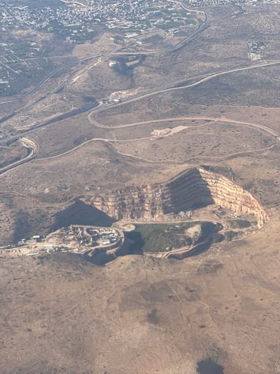 This aerial photograph captures a landscape near Rosh HaAyin, Israel. The scene is dominated by rugged, dry terrain with sparse vegetation. A prominent feature is a large, terraced quarry, its stepped layers carved into the hillside, revealing reddish-brown rock. Adjacent to the quarry, a cluster of buildings and industrial structures suggests ongoing activity, possibly related to mining or processing.

Several roads wind through the landscape. One appears to be a major highway with multiple lanes, curving through the foothills. Other, smaller dirt tracks and paved roads lead to and from the quarry and surrounding areas. On the left side of the image, a densely populated residential area with numerous buildings is visible, contrasting with the more natural, undeveloped land elsewhere. Green patches, possibly agricultural fields or parks, are interspersed within this urban area and along the edges of the undeveloped land.

The lighting suggests it is daytime, with shadows cast by the quarry's terraces and the buildings, indicating the sun is at an angle. The weather appears clear, with no visible cloud cover. The overall impression is one of a semi-arid environment where human development, including infrastructure and industry, interacts with the natural landscape. There is no visible text in the image.