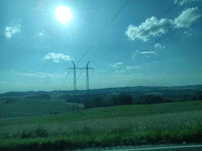 This is an outdoor, daytime photograph taken from a moving vehicle, likely a car or train, looking across a rural landscape. The scene is dominated by a vast, rolling green field in the foreground, with a slightly different texture of grass suggesting it might be a meadow or pasture. In the midground, more fields are visible, some appearing cultivated with darker green crops, and patches of trees and bushes breaking up the open spaces.
Further in the distance, the landscape rolls into gentle hills covered in a mix of forests and possibly scattered settlements. The sky is a bright, clear blue with a few scattered, puffy white clouds. A prominent feature of the sky is the intense glare of the sun, positioned in the upper left quadrant of the image, creating a lens flare effect.
Stretching across the sky are high-voltage power lines, supported by two large, lattice-style metal pylons visible in the midground. These pylons are intricately structured, with the lines originating from them and extending towards the upper right of the frame, disappearing into the distance. The overall impression is one of a peaceful, agricultural region under a bright, sunny sky, with the industrial element of the power lines subtly integrated into the natural scenery. The time of day is clearly daytime, with the strong sunlight indicating it could be late morning or early afternoon. The weather appears to be clear and fair. There is no visible text in the image.