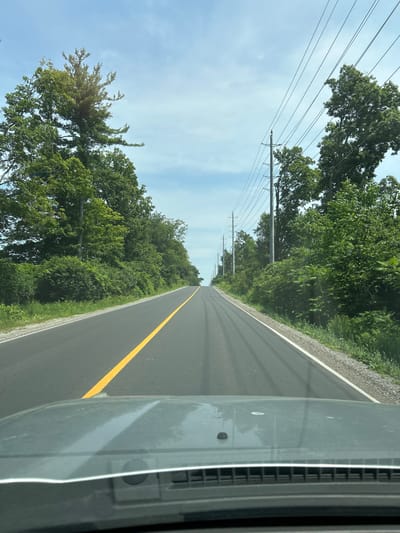 A first-person perspective shot from a vehicle looking forward on a two-lane asphalt road that curves slightly to the left and ascends a gentle hill. A solid yellow line divides the lanes. The right side of the road is lined with green trees and foliage, with power lines and poles visible above the trees. The left side of the road is also lined with trees and foliage. The sky is blue with scattered white clouds. The dashboard and windshield of the vehicle are visible in the foreground, partially obscuring the view of the road. The location is identified as the city of Vaughan, Canada.