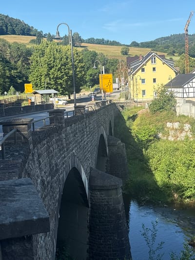 The image captures a scene in Schuld, Germany, focusing on a stone bridge crossing a narrow, flowing river under a clear blue sky. The bridge, constructed of rough-hewn stone blocks, features multiple arched passages. The foreground shows the railing and stone parapet of the bridge.

To the right of the bridge, a steep, grassy embankment is covered in dense green foliage, with some lighter-colored rock formations visible towards the top. Beyond the embankment, a bright yellow building with a dark roof stands prominently, displaying several windows. Adjacent to it, a traditional German half-timbered house with black beams and white infill is partially visible.

Further in the background, rolling green hills covered in trees form the landscape. A construction crane is visible on the right side of the background, indicating ongoing development. In the middle ground, a yellow directional signpost with German text is positioned near the road. A street light with an ornate lamp head stands on the bridge. Several vehicles, including a white van and a car, are present on the road that crosses the bridge. The river water reflects the blue sky. The overall scene depicts a rural or small-town setting in Germany, with a focus on the historic bridge and its surrounding environment.