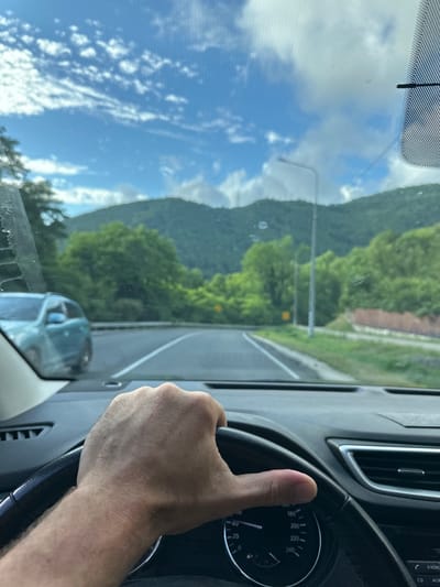 The image is a first-person perspective from inside a car driving on a road. A person's hand is visible gripping the steering wheel. The road ahead is paved with white lane markings and curves slightly to the left. On the right side of the road, there is a grassy embankment with some low vegetation and a stone wall. Beyond the embankment, there are trees and hills covered in lush green foliage. A light pole is visible on the right side of the road. In the distance, mountains are visible under a bright blue sky with scattered white clouds. On the left side of the road, another car, a light blue SUV, is seen driving in the opposite direction. The interior of the car shows a portion of the dashboard with a speedometer and other dials. The overall scene suggests a scenic drive through a mountainous, green landscape, possibly in the area of Volkonka, Russia, given the mention of the location.