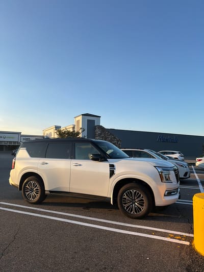A white, two-tone luxury SUV, with black roof and trim, is parked in a shopping center parking lot in East Meadow, New York. The SUV is angled to the right, with its front half closer to the foreground. It has chrome accents on the front grille and wheels. Behind the SUV, the storefronts of various businesses are visible, including "Local Infusion," and a large "Marshalls" store. The sky is a clear, pale blue, indicating it is daytime, likely late afternoon due to the warm light casting long shadows. The parking lot is paved asphalt with white parking lines. A bright yellow parking bumper is visible on the right side of the frame. Other cars are parked in the background, including a white SUV.