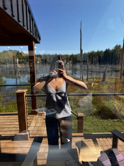 A person is depicted taking a selfie, visible as a reflection in a large glass window overlooking an outdoor scene in Otego, United States. The individual, wearing a light-colored checkered camisole and dark pants, holds a silver smartphone at chest level. A small pendant necklace is visible. The reflection superimposes an exterior view of a calm body of water, likely a lake, dotted with numerous bare, dead trees (snags) protruding from the surface and shoreline. A dense green forest is visible in the background, extending under a clear blue sky. Immediately adjacent to the window, on an outdoor wooden deck with a railing, a wooden table or bench holds a book or magazine. Parts of outdoor chairs are visible at the bottom right, and sunlight casts distinct shadows across the deck's wooden planks.