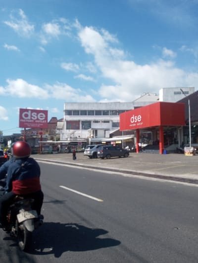 This image depicts a sunlit urban street scene in Ban Phonhxavanh, Laos, characterized by commercial activity under a clear blue sky with scattered white clouds. In the immediate foreground, the back of a person on a motorcycle is visible, wearing a red helmet and a dark blue jacket with a red lower section. The motorcycle casts a distinct shadow on the asphalt road, which features a single white dashed lane marker.

To the right of the road, a large commercial building is present, featuring a prominent red entrance structure emblazoned with "dse FACTORY OUTLET" in white lettering. Two dark-colored cars are parked in an adjacent asphalt lot. Further down the street on the left, another large red billboard-style sign also reads "dse FACTORY OUTLET," accompanied by a "BIG SALE 70%" banner. Beyond these primary structures, additional commercial buildings and street infrastructure are visible. A lone individual is observed walking across the parking area near the parked cars. Other indistinct figures are visible near the building entrances. The overall environment suggests active daytime commerce.
