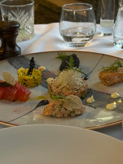 The image shows a plate of food in a restaurant. The plate contains four different dishes. There is a pile of diced salmon on top of chopped greens, a yellow couscous-like dish with a black decorative sprig, a dish of salmon topped with sesame seeds and a green sprig, and a smaller portion of chopped salmon and greens. There is a dollop of white sauce on each of the dishes. The plate is gray and white with black designs on the sides. The table is white, and there are two glasses in the background, one with water and one with an unknown beverage. The image appears to have been taken in a restaurant in Ouled Jelal, Morocco, located at 31.663° N, 7.872° W.
