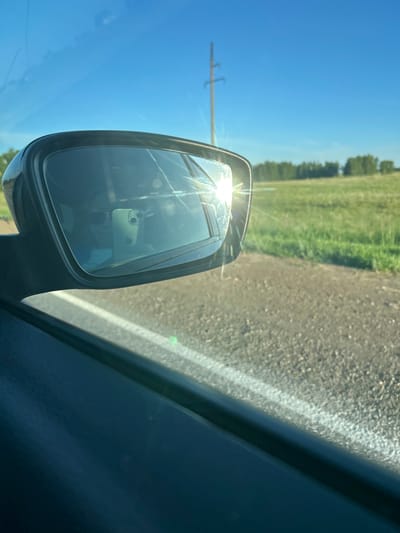 An outdoor, medium shot captures a side-view mirror of a vehicle parked on a dirt road, with a bright sun glare reflecting off the mirror. In the distance, a tree line borders a grassy field, and a utility pole stands against a clear blue sky. The interior of the car is visible in the mirror's reflection, showing a person wearing sunglasses and holding a cell phone. The scene is set in Ishmurzino, Russia.
