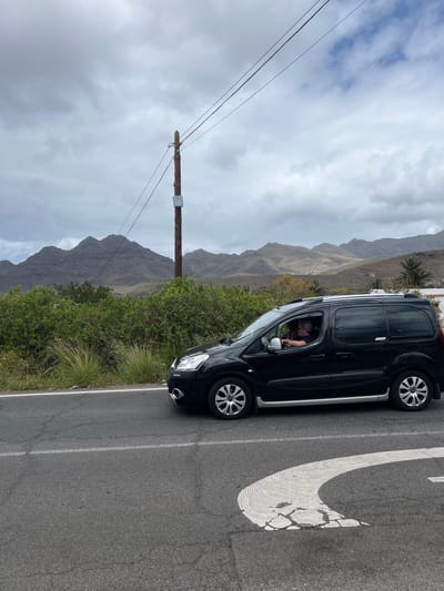The image depicts a black multi-purpose vehicle (MPV) parked on a paved road, likely in La Aldea de San Nicolás, Spain, based on the mountainous landscape and vegetation visible in the background. The road features a worn white directional arrow painted on its surface, indicating a curve or turn. The vehicle is oriented such that the driver's side is facing the camera, and a person is visible inside the vehicle in the driver's seat. Behind the vehicle, a landscape of arid mountains and green shrubbery fills the background. A wooden utility pole with electrical wires extends across the lower portion of the sky. The sky is overcast with grey and white clouds.