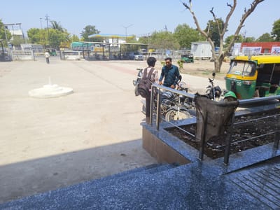 The image presents an outdoor scene in Dhandhuka, India. Two men stand near motorcycles and a railing, engaged in conversation. The man on the left has a backpack. A green and yellow auto-rickshaw is visible to the right of the men. In the background, a man walks away from the viewer and towards what appears to be a bus station or a similar transportation hub, marked by a fenced area and several vehicles. Other buildings, including a petrol station, are also visible. The setting appears to be a public space with concrete flooring and steps leading to a raised platform. A bare tree stands beside the rickshaw. The sky is clear and blue, indicating daylight. The photograph is taken from a slightly elevated position, looking out onto the scene.