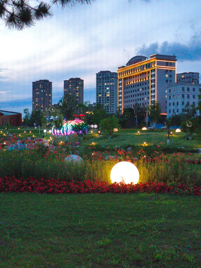 The image depicts a park at dusk with several tall, modern buildings in the background. In the foreground, there is a manicured lawn with a large, glowing white orb light. Behind the lawn, there is a bed of red flowers, and further back, more red flowers and other low-lying plants are visible. Scattered throughout the park are smaller, illuminated spheres and other colorful light installations, including a large, rainbow-colored archway. Several trees are interspersed within the park landscape. The sky is a gradient of blue and gray, with clouds present. The buildings in the background are illuminated, with lights visible in many windows. One building stands out with decorative yellow lighting around its edges and balconies. In the middle ground, there appears to be a large, possibly circular, structure with colorful lights and what looks like a fountain or water feature. Some indistinct figures can be seen in the distance within the park, but they are not clearly identifiable. The overall impression is of an urban park illuminated at night with decorative lighting, set against a backdrop of city architecture.