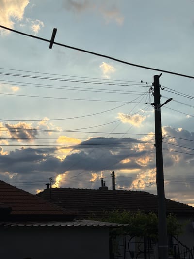 The image depicts a residential area in Dalgopol, Bulgaria, under a dramatic sky. In the foreground, the dark silhouettes of rooftops are visible, suggesting residential buildings. Above them, a complex network of power lines crisscrosses the frame, supported by utility poles. One prominent pole stands on the right side of the image. The sky is filled with a mix of dark, heavy clouds and lighter, illuminated clouds, indicating either an approaching storm or the aftermath of one, with sunlight breaking through. The overall mood is atmospheric and somewhat somber, with the man-made structures contrasting with the powerful natural display in the sky.
