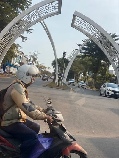 A man wearing a white helmet and a multi-colored jacket rides a motorcycle on a paved road. He is in the foreground, facing right, with his hands on the handlebars. A purple plastic bag is visible on the front of the motorcycle. The road is flanked by trees and buildings on the left, and a modern white archway structure with intricate metalwork on both sides. Several cars are visible on the road in the background, suggesting an urban or suburban environment. The sky is clear and light. The location is identified as Mandar Sari, Indonesia, with a sign visible on the left indicating "MARKETING GALLERY".