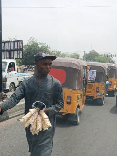 The image displays an outdoor street scene in Hotoro, Nigeria, under a hazy sky. In the foreground, a young male, appearing to be in his late teens or early twenties, walks towards the left side of the frame. He wears a dark baseball cap with light-colored text, a dark-striped long-sleeved tunic, and dark trousers or similar lower garment. His right hand holds a bundle of approximately ten small, clear plastic bags containing a white, popcorn-like substance, along with several smaller, darker-colored bundled items. He looks slightly to his right, away from the viewer.
On the asphalt road behind him, a queue of three yellow and brown three-wheeled auto-rickshaws, commonly known as Keke Napep, are stationary, facing away from the camera. The closest auto-rickshaw has a promotional poster or sticker on its rear displaying an individual and text including "Data App." Further in the background, to the left, a white pickup truck with the marking "DOL-90" on its rear and a blue cargo bed is visible. An individual in a red shirt is seated in the driver's seat of the pickup truck. Green trees and bushes line the mid-ground, and a sign with the text "AN-NOOR" is visible above the auto-rickshaws. A concrete barrier separates the foreground from the vehicle lane.