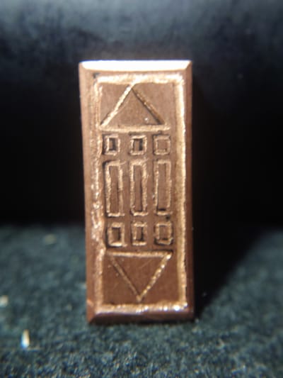 A rectangular copper object with a raised design sits on a dark, textured surface. The design on the object appears to be a stylized representation of a building or structure. The top of the object features a triangle within a square, with smaller squares and rectangles below it, arranged symmetrically. The bottom of the object has an inverted triangle. The lighting in the image highlights the metallic sheen of the object and casts a shadow to its right. The background is dark and out of focus. There is no discernible location information within the image that would suggest it is in Sokolohirsk, Ukraine.