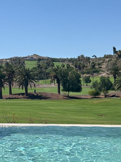 A daytime, eye-level, full shot captures an infinity pool in the foreground, its turquoise water rippling under the bright sun. Beyond the pool, a lush green golf course unfolds, dotted with numerous palm trees and other vegetation. In the distance, a hilly landscape is visible, with a building situated on the highest point. The sky above is a clear, vibrant blue. The scene appears to be located in Urbanización Salobre Golf, Spain, given the characteristic landscape of the area. There are no people visible in the image.