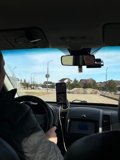 The image captures a first-person perspective from within a vehicle, looking forward through the windshield onto a residential street in Bezborodovo, Russia. The driver's left shoulder and part of their arm, clad in a dark jacket, are visible on the left, with their right hand resting on the steering wheel. A smartphone, displaying a map or navigation application, is vertically mounted on the central dashboard, connected by a white charging cable. Below the smartphone, a car infotainment screen shows a blue display. The rearview mirror is centrally positioned, reflecting a partial view of a face, likely the driver's. A dashcam is mounted to the right of the rearview mirror.
Outside the vehicle, a paved road stretches ahead under a clear blue sky. Multiple streetlights with dark poles line the road. In the background, several detached residential houses with sloped roofs are visible, interspersed with bare deciduous trees and evergreen shrubs. Piles of dirty, melting snow are present on the ground along the roadside and in grassy areas. Further down the street on the left, a white sedan is stationary, with two human figures standing nearby.