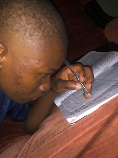 A person lies on a brown plush surface, possibly a couch or bed. The person has dark skin, a shaved head, and acne scarring on their face. They are wearing a dark blue shirt. The person's right hand holds a clear pen above a stack of papers with handwritten text. The background is out of focus, but suggests a residential indoor setting. There is no information in the image to confirm the location of Atani, Nigeria.