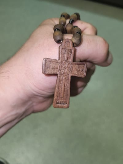 A person's hand is holding a wooden Orthodox Christian cross with a beaded string. The cross is intricately carved with symbols and text, likely religious in nature. The beads are dark brown and appear to be made of wood as well. The background is a plain, light green surface. The scene is a close-up shot, focusing on the cross and hand. The location in the city of Khotsky, Ukraine, is not discernible from the image itself.