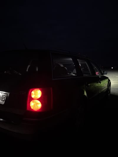 The image depicts a dark-colored car, likely an SUV or hatchback, parked at night in Cherna Gora, Bulgaria. The car is angled diagonally, viewed from a low perspective, showcasing its rear and side. The upper surfaces of the car, including the roof and parts of the side panels, are covered in a textured, greenish-brown material, possibly camouflage, dirt, or moss.
The scene is enveloped in darkness, with the primary light sources being the car's illuminated red taillights, which feature two prominent circular bulbs glowing brightly. A strong, narrow beam of white light shines vertically down onto the upper side of the vehicle, creating a stark contrast and highlighting the car's textured surface against the deep shadows.
Through the rear side window, a glimpse of the car's interior is visible, including seats and a small, pink, possibly plush, object resting on the back seat. Faintly visible on the right taillight is the text "TDI". A portion of the car's license plate is visible at the very bottom, showing the letters "MB". The overall impression is that of a rugged vehicle stopped in a dark, nighttime environment.
