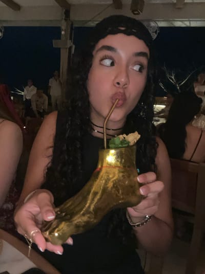 This image depicts a nighttime scene within an outdoor or semi-outdoor restaurant or bar in El Tezal, Mexico. A female subject, positioned centrally, has dark, curly hair and is wearing a black sleeveless top, a black crocheted headband, and multiple black choker necklaces. She is actively drinking from a large, golden, foot-shaped vessel via a bent metal straw. Her lips are pursed around the straw, and her eyes are wide, directed towards the upper right of the frame. The golden vessel contains a liquid, garnished with mint leaves and a disc-shaped item, and a thin red string or ribbon is tied around its upper portion. Her left hand, adorned with rings and featuring French-tipped fingernails, is visible near the vessel. In the blurred background, wooden structural elements are present, along with sparse, tree-like decorative branches and multiple other individuals, some appearing to be staff in white shirts and others patrons.