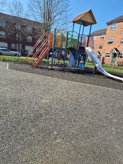The image depicts an unoccupied outdoor children's playground area located within a residential setting in Hereford, United Kingdom, under clear blue skies. In the foreground, a dark, granular safety surface covers the ground. The midground features a multi-colored play structure comprising a raised platform with a light brown peaked roof, accessed by an orange ladder/stairway on the left and featuring a silver slide descending to the ground on the right. The structure's supports are painted in blue, yellow, green, and purple, and it includes various interactive elements such as spherical climbing grips. Behind the playground, residential buildings made of red brick with white-framed windows are visible on both the left and right sides. A black metal fence separates the playground from a driveway or lawn on the left, where a white car is parked. Another fence, partially green, separates the playground from additional residential buildings on the right. A bare deciduous tree stands between the playground and the houses on the left. No individuals are present in the scene.