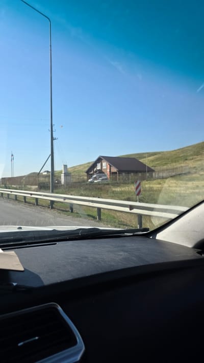 A low-angle, eye-level shot captures a clear, blue sky over a grassy hill in Nizhniy Chiryurt, Russia. In the foreground, a car's dashboard is visible, with a dark texture and a small, light-colored object on the upper left. A black air vent is positioned in the lower left corner.
Beyond the dashboard, a white metal guardrail runs horizontally across the mid-ground, separating the road from the grassy hillside. A tall, gray lamppost stands on the left side of the frame, casting a shadow. A few power lines extend from the lamppost.
On the hillside, a wooden house with a dark roof is visible, surrounded by a wooden fence. Two white vehicles are parked in front of the house. A red and white striped sign is placed on the right side of the frame near the guardrail. Two flags on poles are visible to the left of the lamppost. The overall environment suggests a rural or semi-rural setting under a bright, sunny sky.