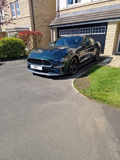 A car with license plate "HM 7" is parked on an asphalt driveway, adjacent to a lawn and a light-colored brick building with a black garage door and a window. The building also has a row of windows on the lower level. A bush with red leaves is positioned next to the car, at the edge of the driveway. This photo was likely taken in Pity Me, United Kingdom.