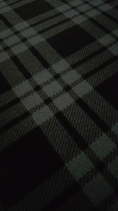 The image is a close-up, overhead shot of a dark plaid blanket. The plaid pattern consists of intersecting dark gray and black lines, creating diamond and rectangular shapes. The texture of the blanket appears to be woven, with a slightly fuzzy finish. The lighting is dim, casting shadows that obscure some of the details of the pattern and the fabric. There are no people, actions, or interactions depicted in the image. The location of Sannikovo, Russia, is not visible or inferable from the image itself.