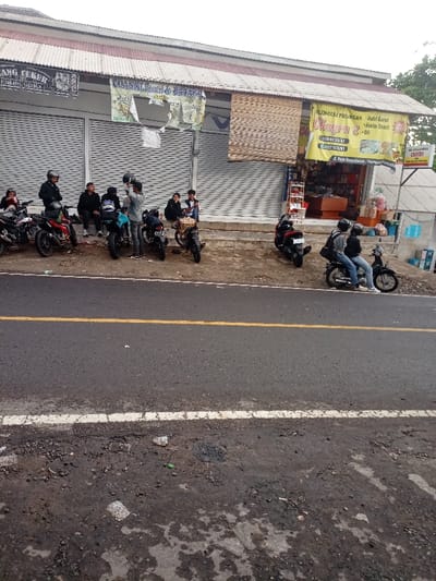 The scene is located at latitude -7.523 and longitude 107.485 in Kecamatan Caringin, Indonesia. It is a street lined with a small, single-story commercial building with a striped awning. The building has roll-up metal doors that are partially open. There are several signs on the exterior of the building, including one for "OLAHGERAK PRIYANGAN - Sate Goat - Ayam Betutu" and "WARUNG 2" which appear to be food vendors. Seven individuals, all men, are standing in front of the building. Some are talking, others are looking at their phones. There are seven motorcycles parked in front of the building. The motorcycles are all facing the street, and some are still running. Two men are sitting on one of the motorcycles, wearing helmets and looking to the right of the scene. The pavement is dark grey and asphalt with a white stripe down the center, indicating a single lane of traffic.