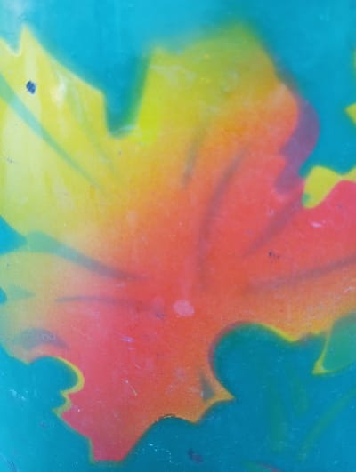 The image displays a close-up, abstract view of spray-painted art on a vertical surface. The dominant colors are teal, yellow, and orange, blended together in a gradient effect that suggests a stylized depiction of a leaf or floral pattern. The texture appears slightly rough, consistent with spray paint application. There are no people, discernible objects, or actions depicted. The location in Voznesenske, Ukraine is not visually ascertainable from the image itself.