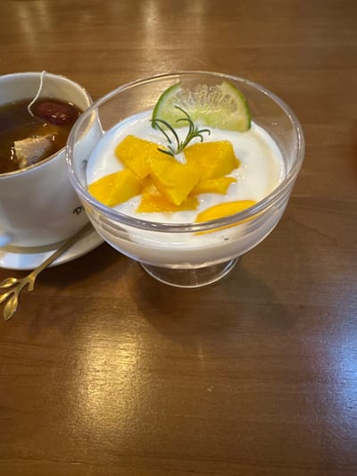 The image displays a close-up, overhead view of a dessert and beverage situated on a dark brown wooden table in Buôn Ma Thuột, Vietnam. A clear glass dish, positioned centrally, contains a white, creamy substance, likely a yogurt or pudding, adorned with numerous irregular diced pieces of bright yellow mango, a thin green lime wedge, and a small sprig of green herb (possibly rosemary). To the left, partially cropped, a white ceramic cup with a gold rim and faint dark lettering contains a dark amber liquid, presumed to be tea, with a white tea bag string visible over the rim, along with a reddish-brown fruit (possibly a dried jujube) and another lighter ingredient submerged. This cup rests on a white ceramic saucer, also featuring a gold rim, on which a small, gold-colored metallic spoon with an ornate leaf-shaped handle is placed. The wooden table surface exhibits a visible grain pattern and reflects overhead lighting. No individuals are present, and no active interactions or actions are depicted within the scene.