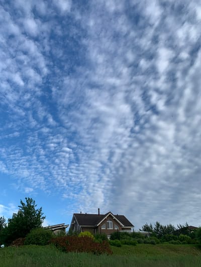 The image captures a wide-angle view of a partly cloudy sky with a bright blue expanse and scattered white clouds, some appearing as streaks and others as smaller, rounded formations. The perspective is tilted, with the sky dominating the left half of the frame. On the right side of the image, a portion of a residential area is visible, featuring the roofs and upper floors of two houses. The houses appear to be modern, constructed with brick and dark roofing tiles. Lush green grass and trees form a border along the right edge of the frame, suggesting an outdoor setting in a suburban or semi-rural environment. One of the houses has prominent gabled roofs and visible windows. The lighting suggests it is daytime, with the sun likely high in the sky given the brightness of the clouds and blue sky. The overall atmosphere is serene and tranquil. There is no visible text in the image.