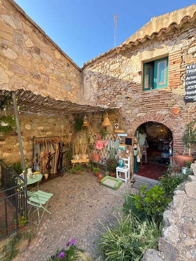 The image depicts an outdoor courtyard area in Tossa de Mar, Spain, likely a small shop or artisan studio. The courtyard is paved with cobblestones and surrounded by stone walls, some of which are reinforced with brick. A covered area, with a roof made of woven reeds or straw, provides shade over a section of the courtyard.
To the left, a metal railing encloses a descending staircase, with potted plants and a small green metal table and chairs set up. Clothing items are displayed on a rolling rack under the reed covering. Behind the clothing rack, a small wooden bench holds decorative ceramic vases.
To the right, an arched doorway leads into the interior of a shop, where more clothing and items are visible. A small white chair sits in front of the shop entrance, next to a display of handbags hanging on a white trellis. Various potted plants and greenery are scattered throughout the courtyard and around the shop entrance, adding to the rustic ambiance. A black sign with white lettering is mounted on the wall near the shop entrance, appearing to list shop offerings. The sky above is clear and blue, suggesting fair weather. The overall scene conveys a quaint, artisanal atmosphere typical of a Mediterranean coastal town.