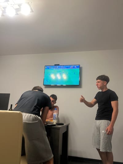 In a dimly lit room, three young men are gathered around a television, engrossed in watching a soccer match. The room appears to be a living space, with a light-colored wall and a large screen television mounted on it, displaying the game. To the left, a bright, multi-bulb lamp illuminates part of the wall.

One of the men, wearing a black t-shirt and gray shorts, stands near the television, pointing at the screen with a curious expression. Another man, also in a black t-shirt, is seated, leaning back and looking at the television. A third man is seated in the foreground, his back mostly to the camera, also watching the game. A dark wooden furniture unit is visible beneath the television, holding a bottle of water with visible text reading "OPE KONA 37". The presence of a bottle and what appears to be a snack on the furniture suggests a casual viewing experience.

The lighting suggests it could be evening or nighttime. The focus is clearly on the shared activity of watching the soccer game, with the men appearing engaged and focused on the screen. The setting seems to be a modern indoor space, possibly a home in Chaidefto, Greece, given the location context.