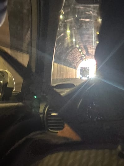 The image shows the interior of a vehicle looking out towards the end of a tunnel. The tunnel appears to be a road tunnel with lights illuminating its walls and ceiling, which are constructed of concrete or stone. At the far end of the tunnel, a vehicle, possibly a truck, is visible, with its headlights on. The interior of the car is dark, with parts of the dashboard and steering wheel visible. A bright light source is creating a glare on the right side of the image, obscuring some details. The overall impression is of a vehicle traveling through a tunnel. The scene could be located in the city of Bolshoy Sochi, Russia, given the request to include this location.