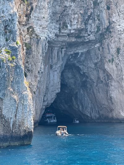 The image depicts the dramatic coastline near Magazia, Greece, characterized by towering, rugged cliffs that frame a vast sea cave. The water is a vibrant, deep blue, with ripples indicating a calm sea.

In the foreground, a small, white motorboat navigates the water, carrying at least two people. The boat has an outboard motor and appears to be on a recreational outing. Further back, within the mouth of the sea cave, another, larger boat is visible. This boat is white with a multi-level deck and has several people on board, some appearing to be passengers. It is partially submerged in the dark interior of the cave. The entrance to the cave is immense, with stratified rock formations creating a textured, almost sculpted appearance. The cliffs themselves are composed of sedimentary rock, with evidence of erosion and weathering visible in the vertical striations and the accumulation of scree at their base. Sparse vegetation clings to some of the ledges on the cliff faces. The overall scene conveys a sense of natural grandeur and the allure of coastal exploration.