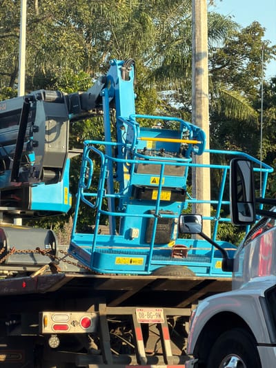 A blue boom lift sits atop a flatbed truck, positioned against a backdrop of trees and sky. The lift features a caged platform, a control panel, and hydraulic components. The truck includes a partially visible silver cab, a license plate (M6-98-80), and various lights. Chains are visible, securing the lift.