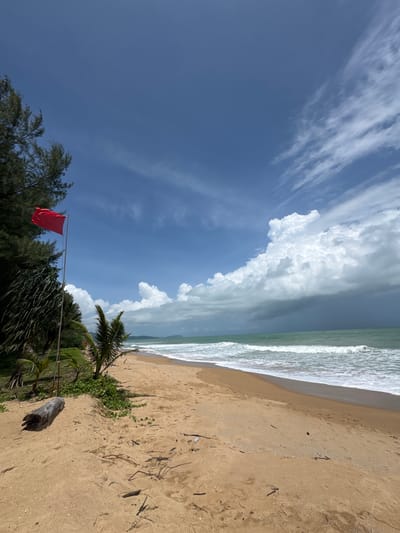 A wide-angle, low-angle shot captures a tropical beach scene in Ban Khuk Khak, Thailand. A bright red flag flies from a pole on the left side of the frame, near a cluster of dark green trees and palm fronds. The sand in the foreground is light brown, with some scattered driftwood and debris. Gentle waves roll onto the shore, with the ocean stretching towards the horizon. The sky above is a vibrant blue, dotted with white, fluffy clouds and wispy cirrus formations. In the distance, the coastline is visible, with more trees and landmasses. The overall impression is one of a serene, natural environment, though the red flag suggests a warning or caution.