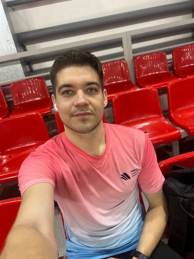 A medium close-up shot captures a man taking a selfie while seated in what appears to be a stadium or arena. The image is oriented sideways, with the man's head on the left and his body extending to the right.
The man has short, neatly styled dark brown hair and light blue eyes. He has light stubble on his chin and upper lip. His expression is neutral, looking directly at the camera. He is wearing white wireless earbuds in both ears.
He is dressed in a short-sleeved athletic t-shirt that features an ombre design, transitioning from a vibrant coral pink at the top to a light blue at the bottom. On the left side of his chest, a black logo consisting of three curved, horizontal lines is visible, along with the text "Gadove Vodovod Marathon". A black digital watch is visible on his left wrist.
The background consists of rows of empty, bright red plastic seats, typical of a sports venue or auditorium. Behind and to the left of the man, there's a light gray metal structure, possibly part of the seating support or a barrier. A black bag is partially visible to the right of the man's shoulder, resting on an adjacent seat. The lighting suggests an indoor setting, possibly artificial lighting.