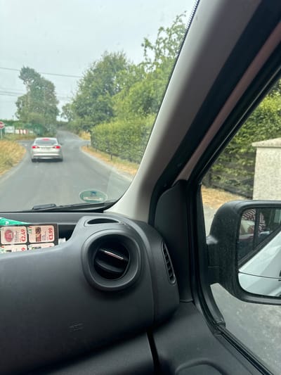 An external, slightly hazy, eye-level, medium shot captures a view from inside a vehicle looking out at a two-lane asphalt road. The road curves to the left and is lined with green foliage and trees. A silver sedan is driving away from the camera on the road. The sky is overcast and grey. On the dashboard in the foreground, there are two product advertisements with prices displayed, an air vent, and an airbag warning label. The side mirror of the vehicle is visible on the right, reflecting a red and white patterned object, likely clothing. The image appears to be taken in Bamford Cross, Ireland, based on the context provided.