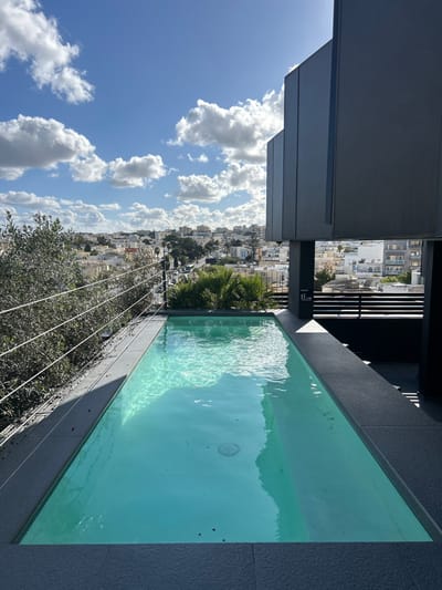 The image displays an elevated perspective of Saint Julian's, Malta, featuring a narrow, rectangular plunge pool in the foreground. The pool contains clear, turquoise water, reflecting a bright blue sky with scattered white cumulus clouds. Dark gray textured tiles surround the pool. Along the left edge of the pool deck, a safety railing composed of horizontal wires is visible, beyond which dense green foliage of various trees and shrubs extends downwards. To the immediate right of the pool, a multi-faceted modern building structure, clad in dark gray panels, rises vertically. A sign indicating "P: 1.10m" is affixed to a pillar adjacent to the pool edge on the right. In the middle ground and background, a sprawling urban landscape of Saint Julian's unfolds, characterized by numerous light-colored buildings, a visible road with vehicular traffic, and additional patches of greenery. No individuals are present in the scene.