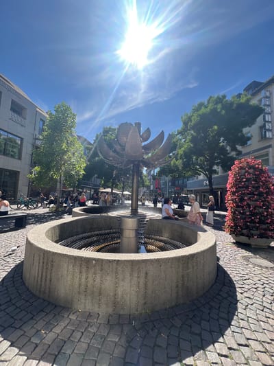 This is an outdoor shot of a fountain in a city, likely Aachen, Germany, on a sunny day. The sun is bright and positioned in the upper left of the frame, casting long shadows across the cobblestone pavement.
The main subject is a circular, concrete fountain with metal elements. Inside the fountain, there are multiple tiers of what appear to be small, round objects, possibly beads or stones, creating a textured surface. A metallic pipe extends upwards from the center of the fountain.
Several people are visible around the fountain and in the background. Some are sitting on the edge of the fountain, while others are standing or walking nearby. One woman, wearing a pink dress, is seen interacting with the fountain. In the background, there are buildings, trees, and shops lining a street. The presence of parked bicycles suggests a public urban space.
The time of day appears to be midday or early afternoon, given the bright sun and the length of the shadows. The weather is clearly sunny and pleasant. The overall mood is relaxed and social, with people enjoying the outdoor space.
There is some visible text on the buildings in the background, though it is mostly illegible due to the distance and image resolution. One sign appears to have the letters "CH" and "EN" visible. Another sign has "WIR" in red letters.
