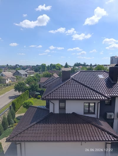This media file captures an aerial view of a suburban neighborhood on a clear, sunny day. The scene is dominated by a collection of houses with dark brown tiled roofs, a common architectural feature in the area. In the foreground, a modern house with white and dark gray siding is visible, featuring several windows and an outdoor air conditioning unit. Its prominent brown roof extends across the frame. Adjacent to this house, a neatly manicured lawn is bordered by a tall, dark green hedge and a stone wall, with a paved pathway leading to the street.
Beyond the immediate foreground, the image reveals a street lined with more houses, many of which also have tiled roofs and are surrounded by lush green trees and vegetation. In the distance, a dense line of trees forms the horizon, behind which are glimpses of other buildings and what appear to be church steeples, suggesting a populated area. The sky above is a vibrant blue, dotted with fluffy white clouds, indicative of fair weather. The lighting suggests it is midday, with the sun casting shadows on the roofs and lawns.
The overall impression is one of a peaceful, well-maintained residential area in Novovolynsk, Ukraine. There are no people visible in the frame, and the focus is on the architecture and the natural surroundings. The timestamp "26.06.2026 12:22" is superimposed on the bottom right of the image.