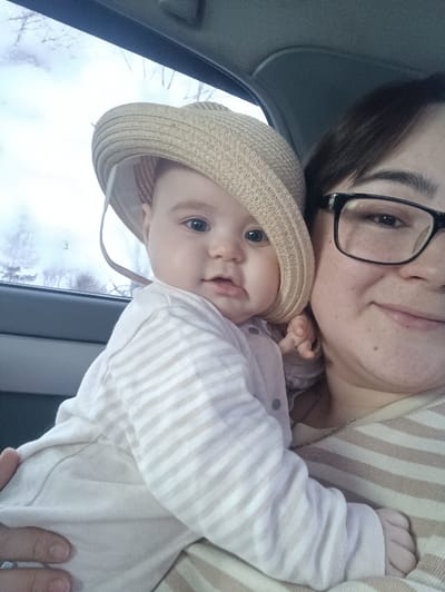 The image displays an adult female holding an infant within the passenger compartment of a vehicle. The adult, positioned on the right, presents a partial view of her face, characterized by dark hair, dark-framed glasses, and a slight smile. She wears a beige and white horizontally striped garment. The infant, centered in the frame and held by the adult, wears a light-colored long-sleeved top with thin horizontal beige stripes and a light beige straw hat with a wide brim, partially obscuring the infant's head. The infant has light skin, blue eyes, and looks directly towards the camera. The background, visible through the vehicle window, is blurred, showing what appears to be bare trees under an overcast sky, indicative of a cool climate. No specific geographical identifiers within the image confirm the location as Simakovo, Russia.