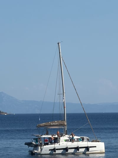 A white catamaran sailboat, identified by the text "BALI 4.1" on its hull and sail, is anchored in clear, calm blue water. The boat has two hulls and a mast. Three men, appearing to be in casual attire, are on the deck of the sailboat. One man is standing near the front of the boat, another is leaning on a railing, and a third is standing on a higher level at the rear. A gray inflatable dinghy is attached to the stern of the catamaran. A Greek flag is flying from the mast. In the background, a range of hazy, mountainous landmasses is visible against a clear, pale blue sky. The scene is characteristic of the coastal areas around Skiathos, Greece, a popular destination for sailing and boating.