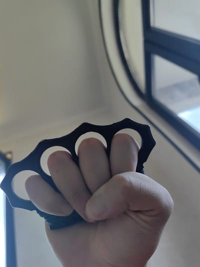 A close-up, low-angle shot shows a person's right hand, clenched into a fist, wearing a set of four black brass knuckles. The knuckles are positioned over the index, middle, ring, and pinky fingers. The background is out of focus and appears to be an indoor environment with a window frame visible on the right. There are no discernible elements in the image that would indicate a specific location within the city of Ingiri, Georgia.